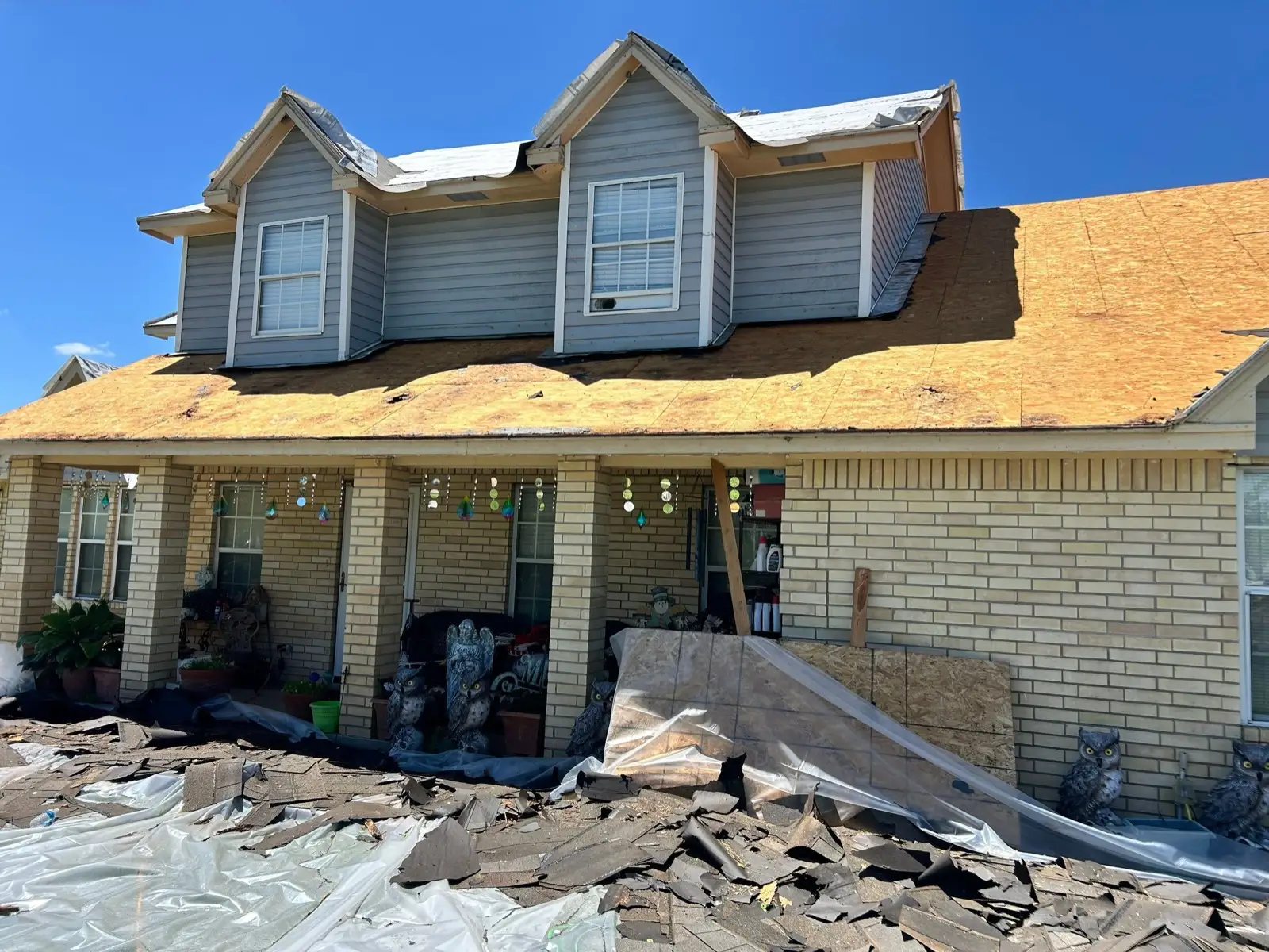 May 14. Two layers gone — bare decking overhead, the old shingles heaped on sheeting below, the porch boarded and tarped.
